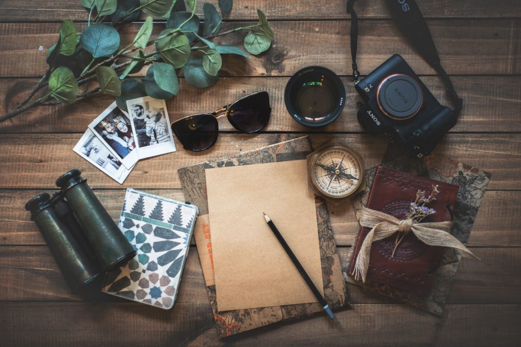A table with a journal, compass, binoculars, a camera, and sunglasses on it.