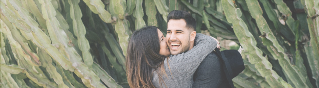 woman hugging man and kissing him on the cheek in front of green cacti