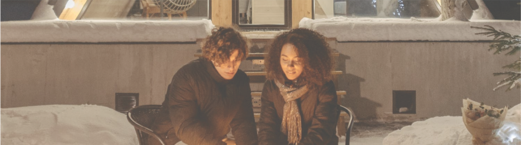 Couple warming hands by the fire in front of cabin surrounded by snow
